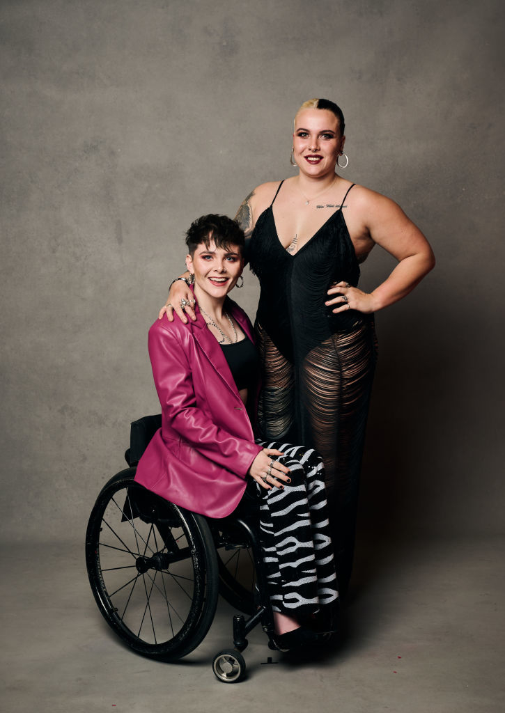Lauren in a wheelchair wearing bright blazer and zebra pants, and Jude in a dark bodysuit with ripped tights, they're posing with arms around each other