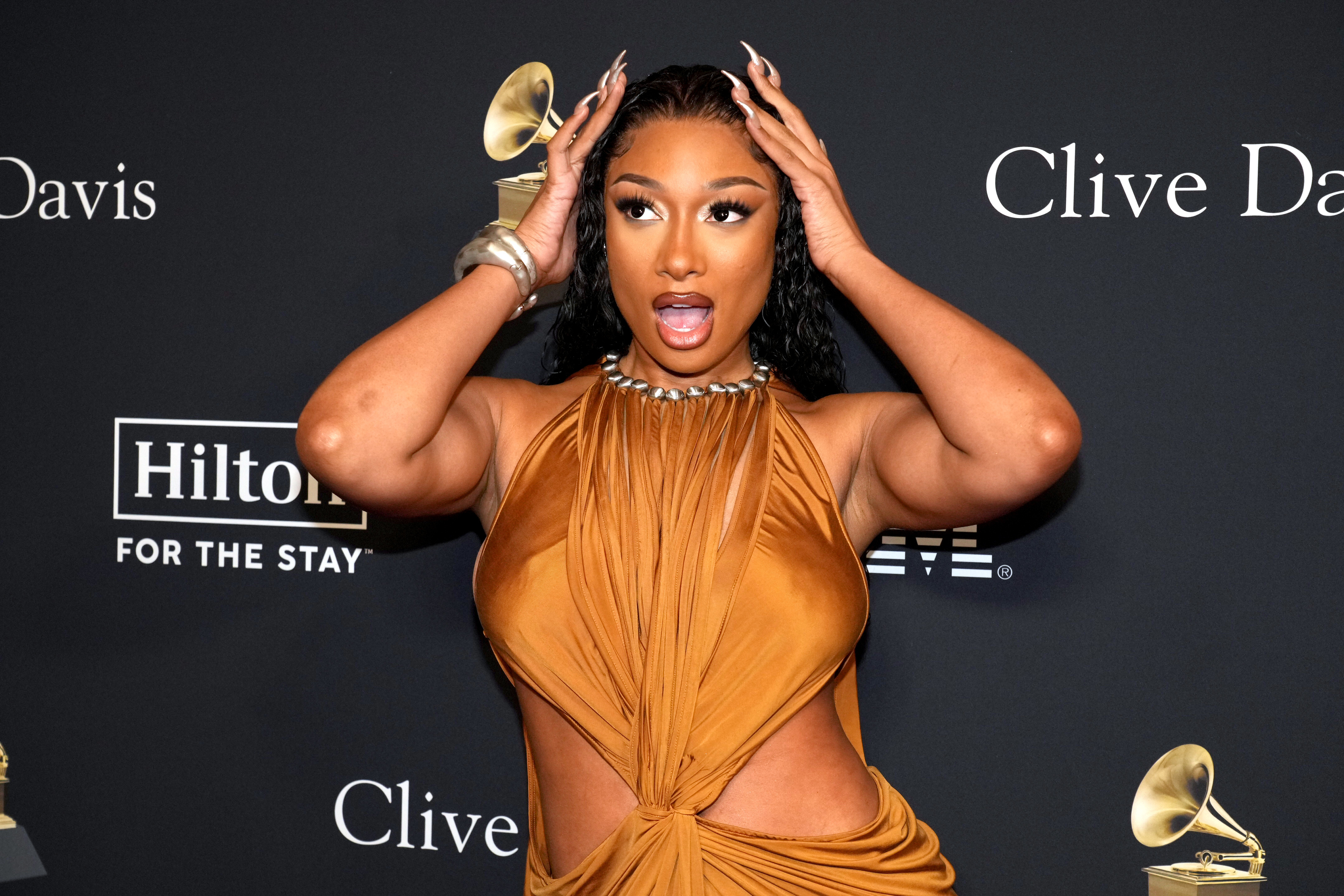 Megan Thee Stallion poses with her hands on her head, wearing a stylish draped dress at an event with Clive Davis and Grammy logos in the background