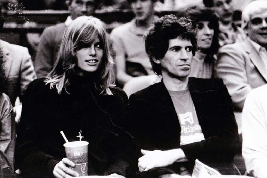 Anita Pallenberg and Keith Richards sitting together at an event. She is holding a drink cup while they both look ahead. Other people are visible in the background