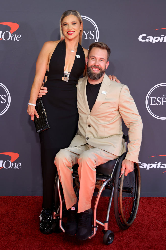 Oksana and Aaron with their arms around each other on a red carpet, her standing in a dress with clutch and him in a wheelchair in a light suit