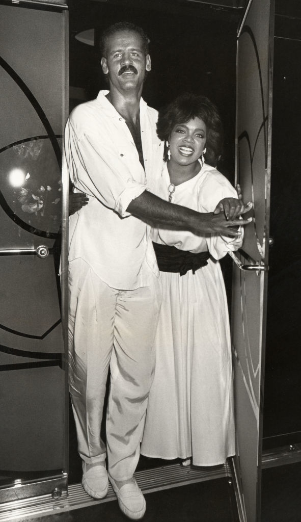Reggie Jackson and Oprah Winfrey walk through a door, smiling and holding matching outfits, white shirts and pants for Reggie, and white dress for Oprah