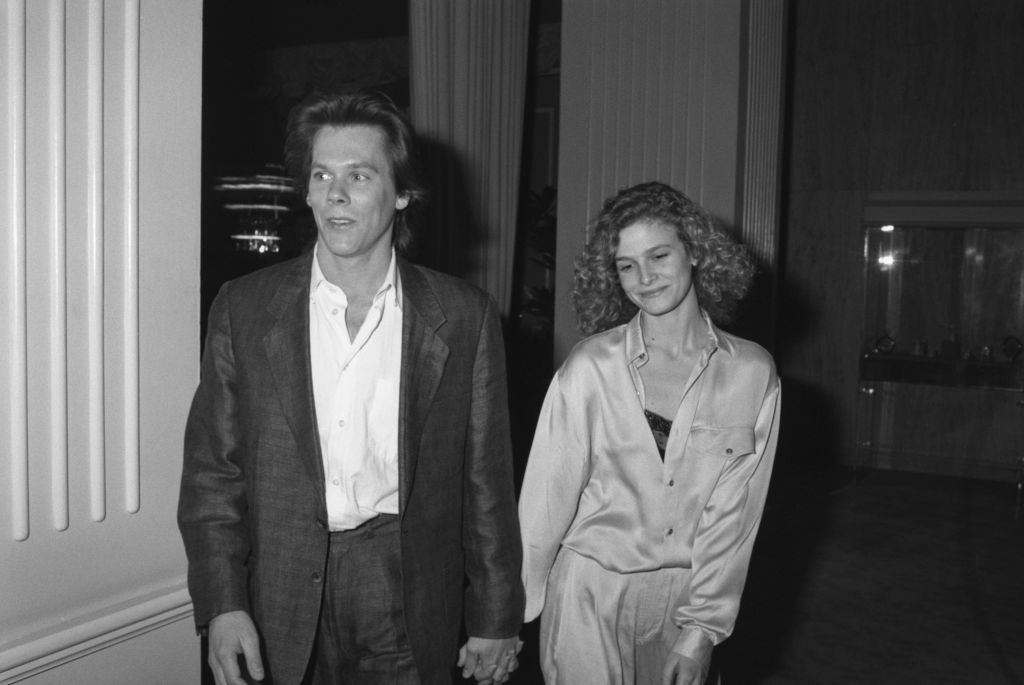 Kevin Bacon and Kyra Sedgwick walking hand in hand. Kevin is in a casual suit, and Kyra is in a loose shirt and pants