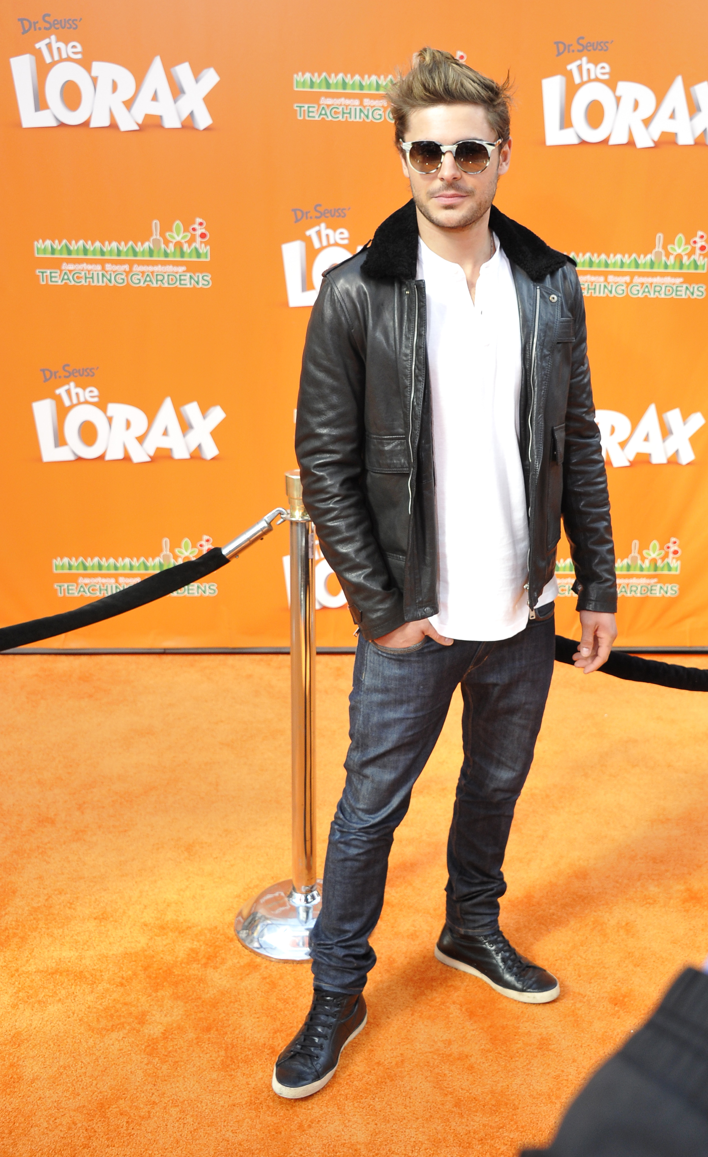 Zac Efron stands on the red carpet at "The Lorax" event, wearing sunglasses, a black leather jacket with a white shirt, blue jeans, and black sneakers