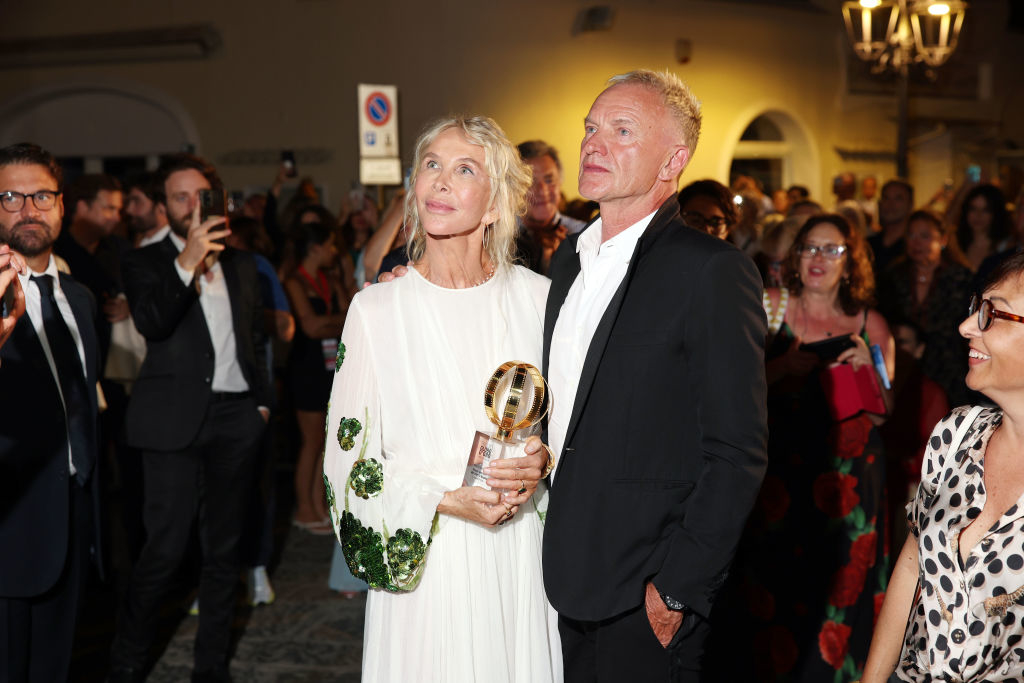 Trudie Styler and Sting at an event, with Trudie holding an award. They are surrounded by other attendees in semi-formal attire