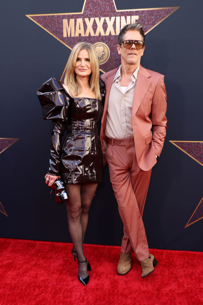 Kyra Sedgwick in a metallic dress with exaggerated sleeves, and Kevin Bacon in a light suit with sunglasses on the red carpet in front of the "Maxxxine" backdrop