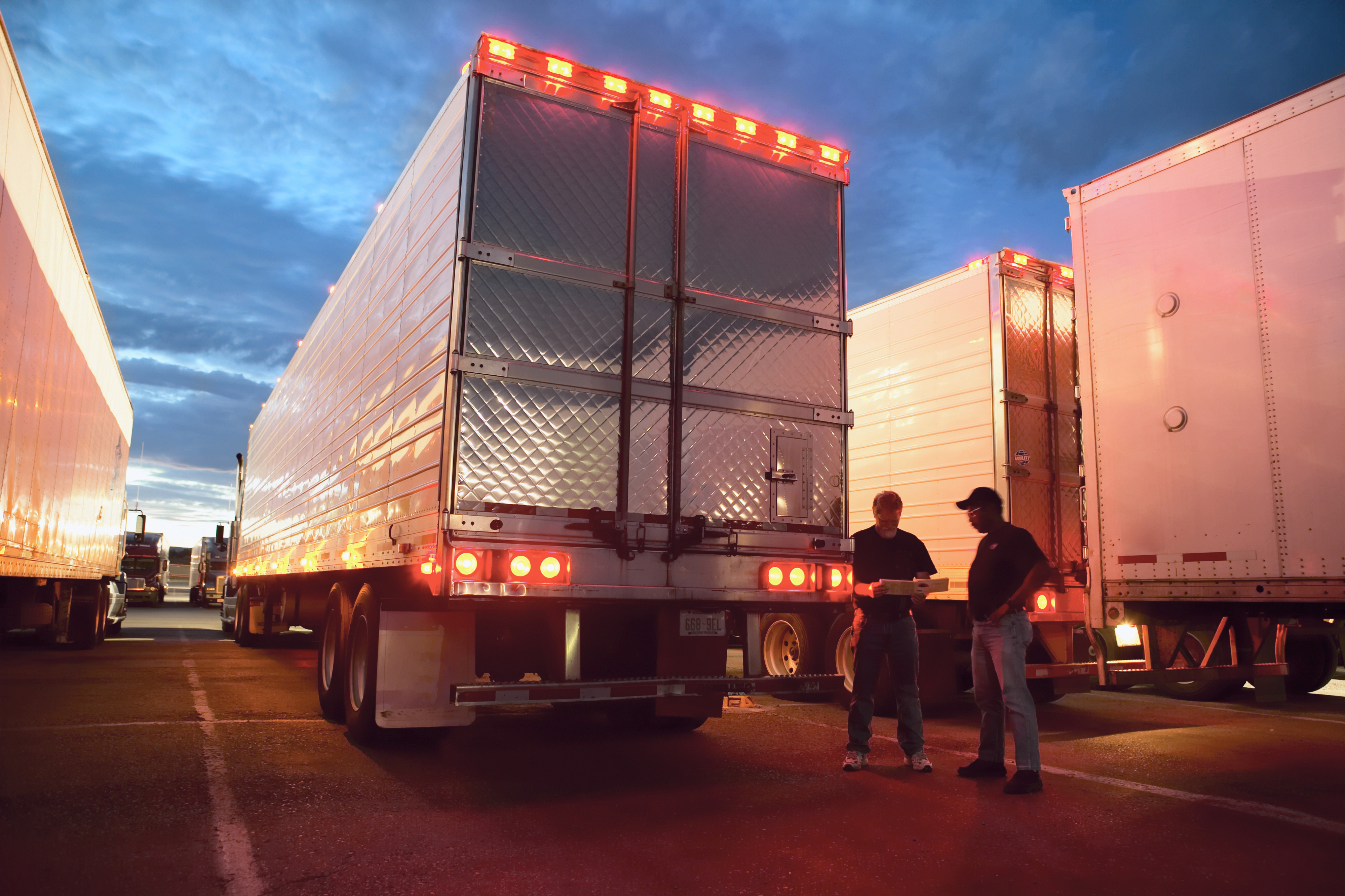 Two people stand between large trucks at dusk, engaged in conversation next to the reflective back of a truck trailer