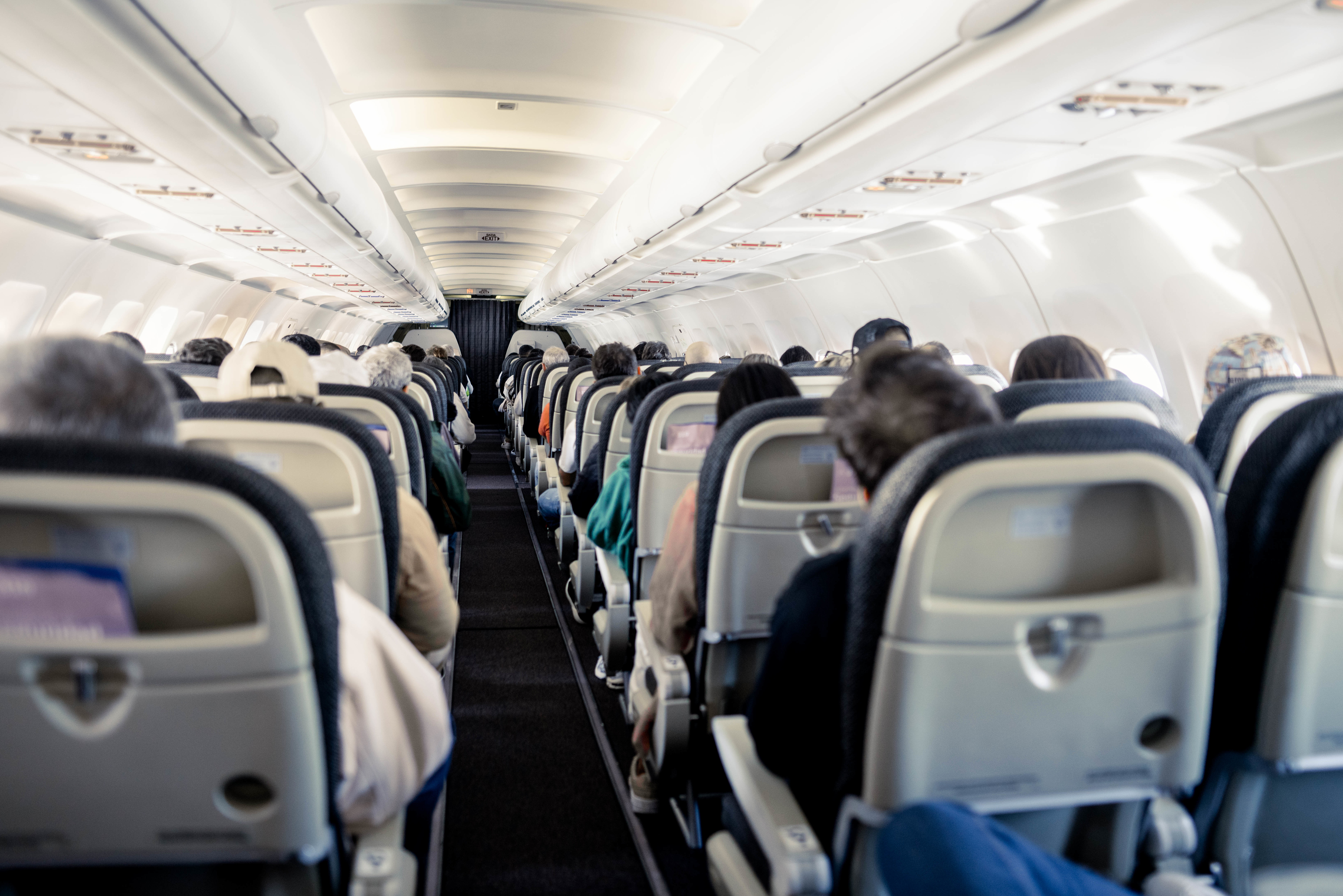Passengers seated inside an airplane cabin, viewed from the back towards the cockpit, focusing on the rows of seats and passengers