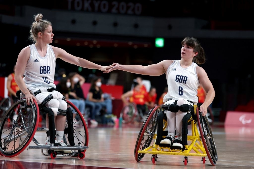 Robyn and Laurie in wheelchairs wearing GBR jerseys, numbered 13 and 8, hold hands on a basketball court during the Tokyo 2020 Paralympics