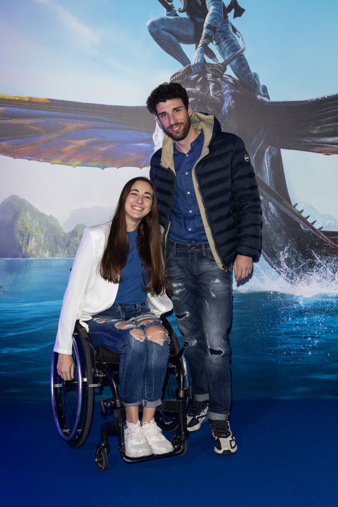 Stefano and Giulia posing together on a red carpet, her in a wheelchair wearing rips jeans and a t-shirt and cardigan and him standing wearing ripped jeans, button-up shirt, and puffer jacket