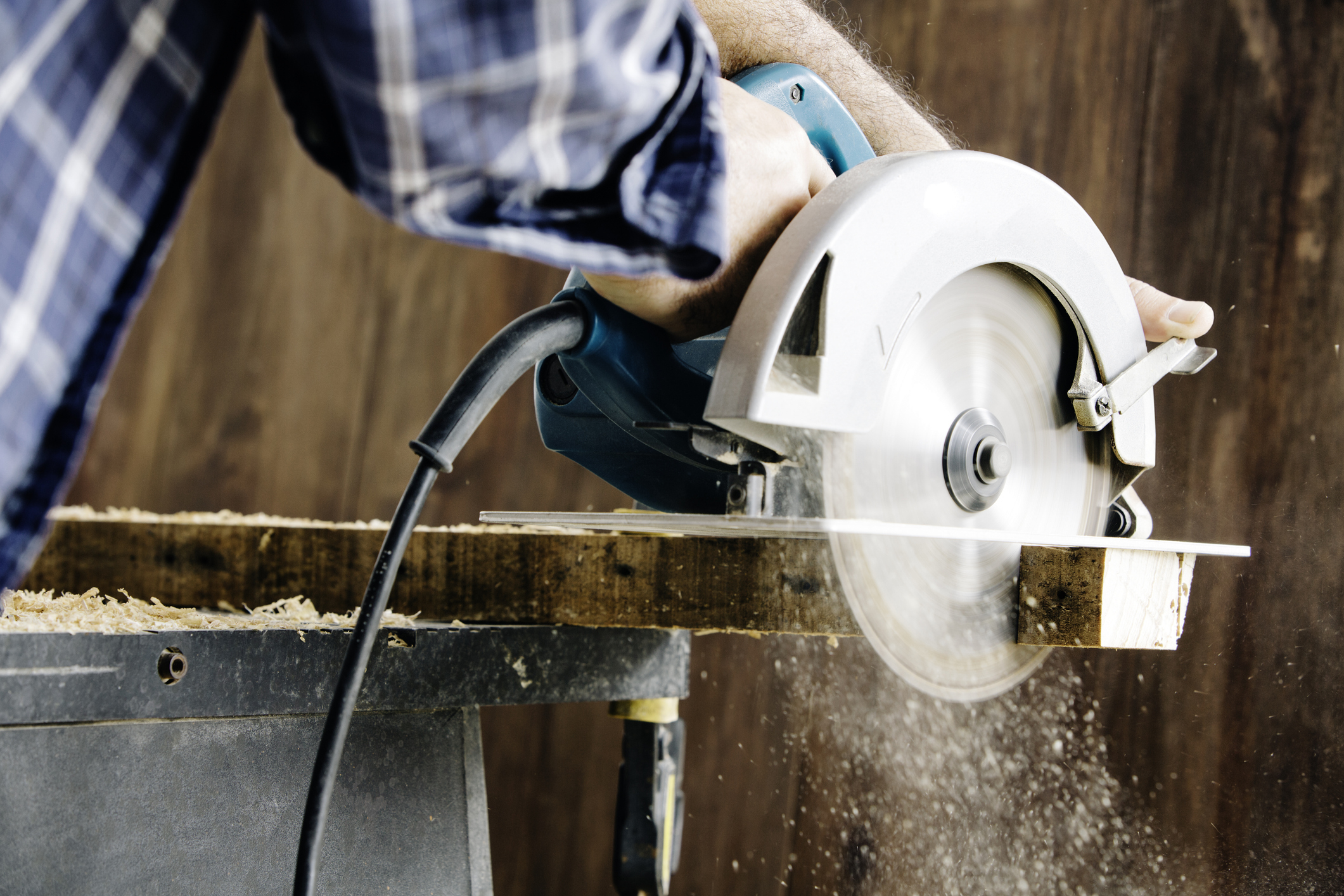 A person in a plaid shirt is using a circular saw to cut a piece of wood, creating sawdust