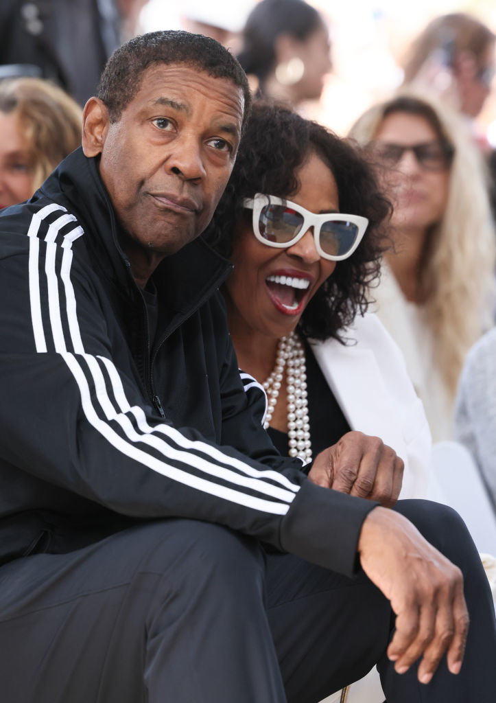 Denzel Washington in a casual tracksuit, seated next to Pauletta Washington, who is wearing a white jacket and sunglasses, both smiling and enjoying an event