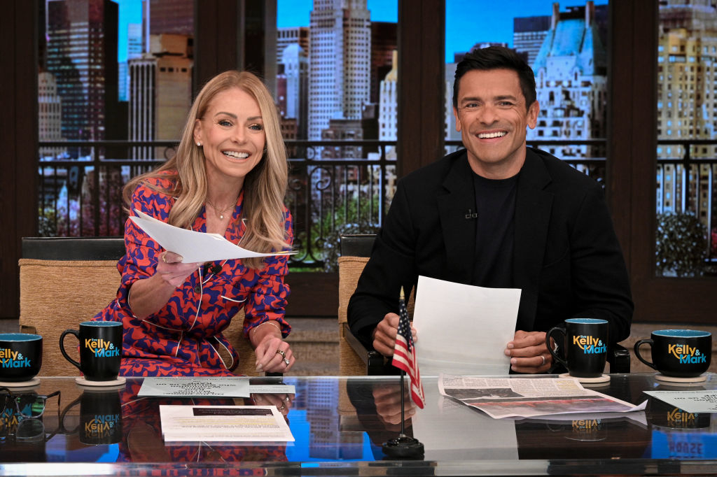 Kelly Ripa and Mark Consuelos sit at a desk on the "Live with Kelly and Mark" set, smiling and holding papers. Both wear formal attire