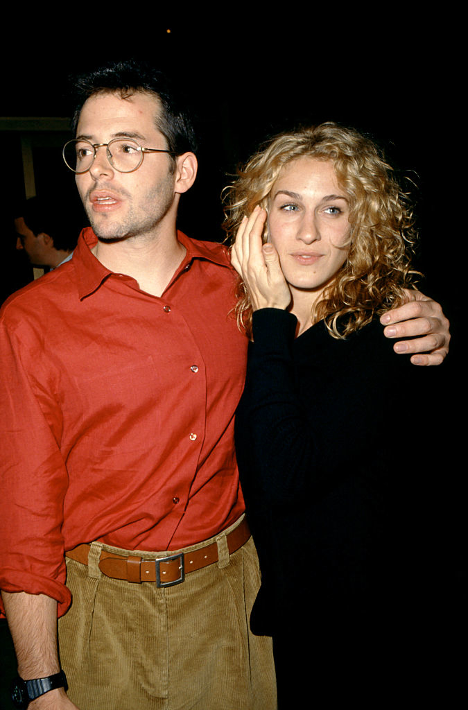 Sarah Jessica Parker and Matthew Broderick pose together. Sarah is wearing a black outfit, while Matthew wears a red shirt and khaki trousers with glasses