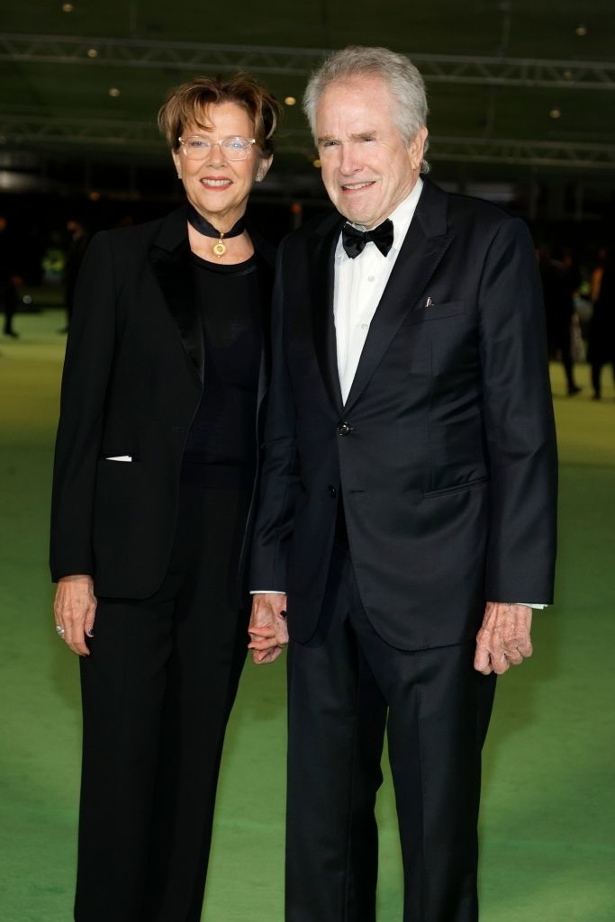 Annette Bening and Warren Beatty smile on the red carpet, both dressed in elegant black formal attire. Annette wears a necklace, and Warren wears a bow tie