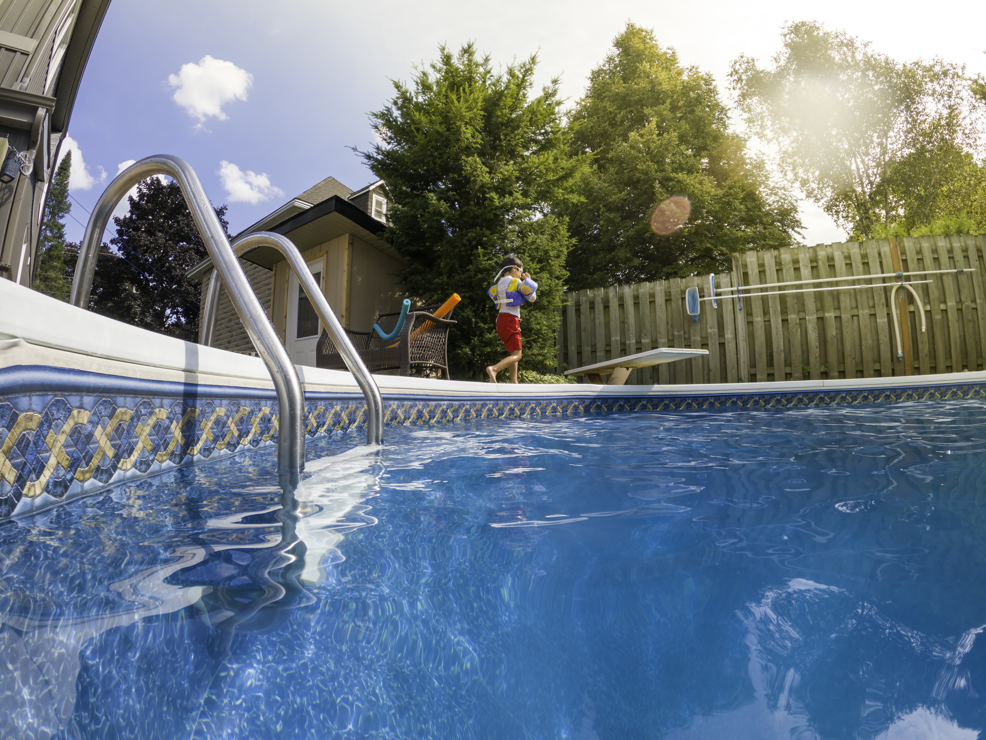 Child in a life vest stands at the edge of a swimming pool, about to jump in. Trees and a house are in the background