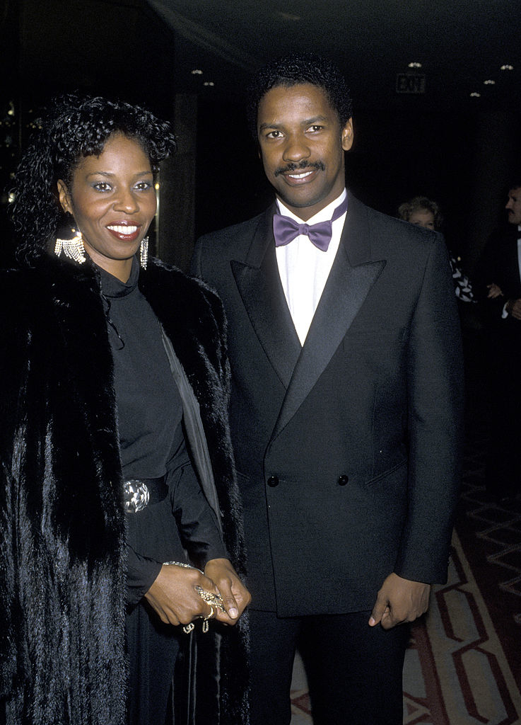 Denzel Washington and Pauletta Washington pose for a photo at an event. Pauletta wears a black outfit with a fur coat; Denzel is in a classic tuxedo with a bow tie