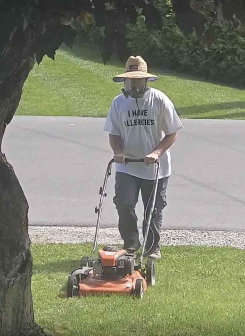 Person mowing a lawn wears a hat, mask, and t-shirt reading "I HAVE ALLERGIES." Tree trunk is visible in the foreground