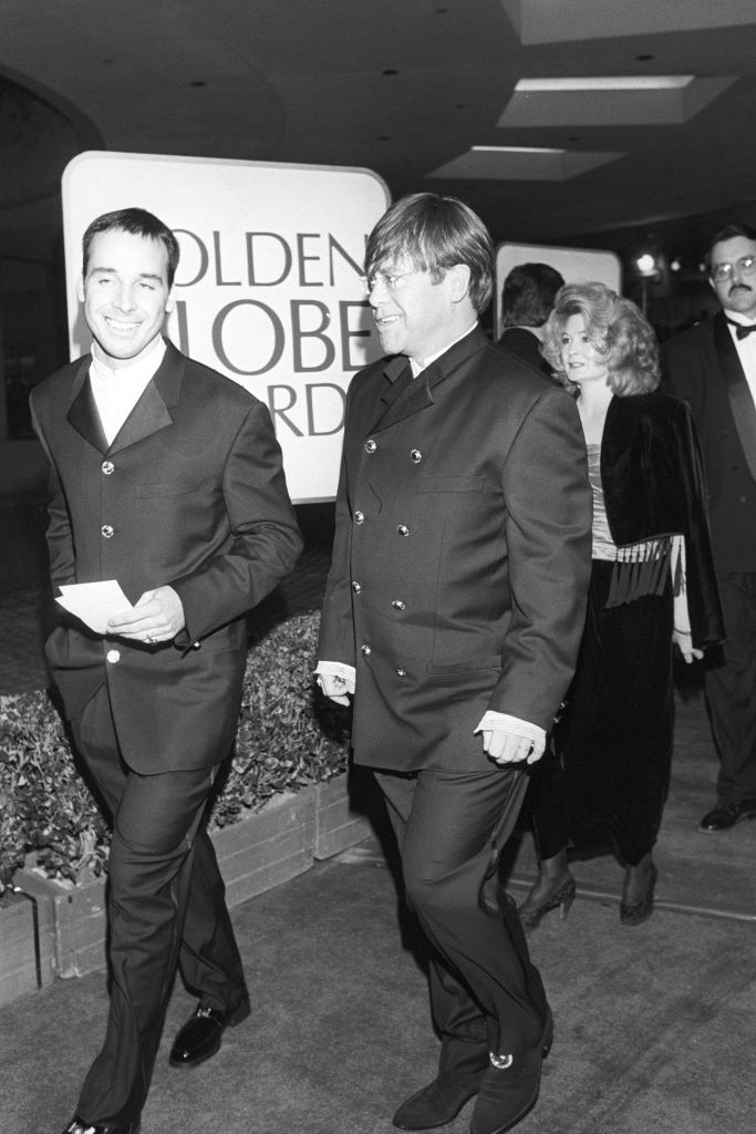 Two men, John Denver and guest, in matching suits, walk the Golden Globe Awards red carpet, followed by a woman in a velvet jacket and a man in a suit