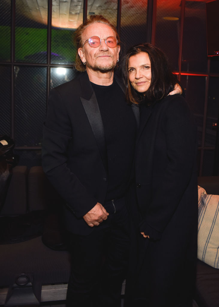 Bono, wearing a black suit and glasses, stands with Ali Hewson, who is wearing a black coat, in a warmly lit indoor setting