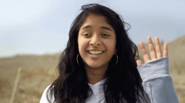 Maitreyi Ramakrishnan smiles while waving her hand outdoors. She has long, wavy hair and is wearing a light sweater