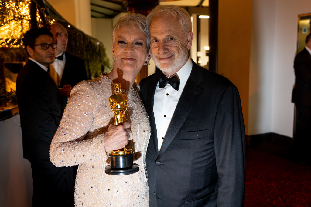 Jamie Lee Curtis, in an elegant embellished gown, holds an Oscar statue while smiling with a man in a classic black tuxedo