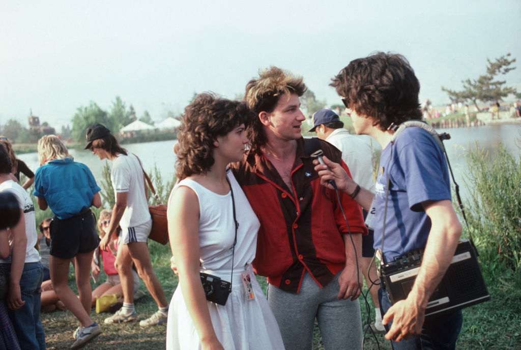 Patrick Swayze being interviewed outdoors at an event; a woman in a white dress stands next to him while the interviewer holds a microphone