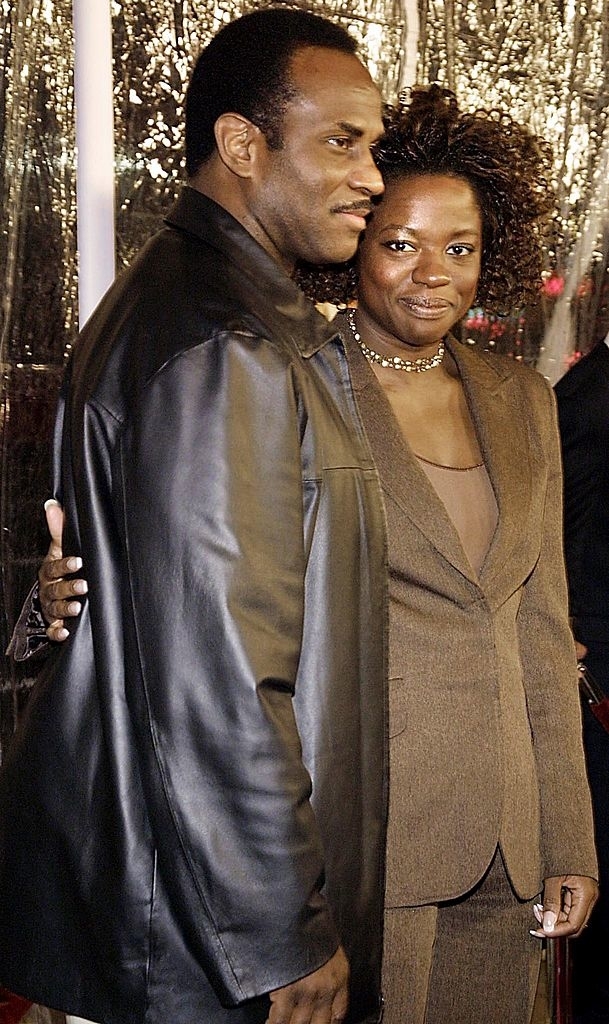A man and Viola Davis pose on a red carpet. The man wears a leather jacket, while Viola Davis is in a blazer