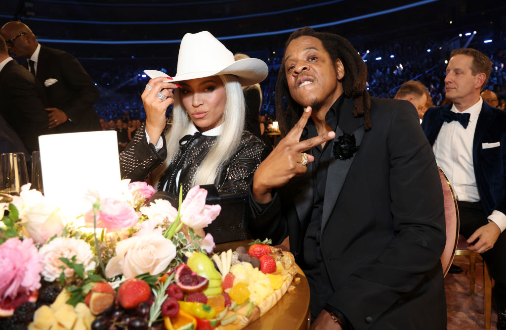 Kim Kardashian in a cowboy hat and glittering outfit, posing with Jay-Z, who is giving a peace sign at an event with a lavish fruit platter in front of them