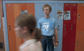A person with curly hair and glasses stands holding books in front of school lockers. Another person walks by quickly, obscuring part of the view