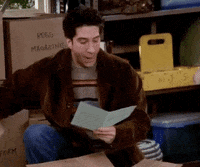 Ross from Friends, wearing a fur coat and striped sweater, enthusiastically reads a card while surrounded by boxes