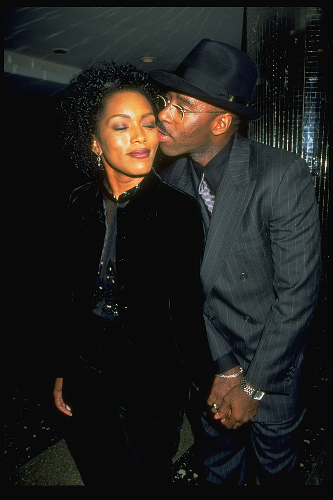 Angela Bassett in a stylish black outfit, receiving a kiss on the cheek from a man in a dark pinstripe suit and hat on the red carpet
