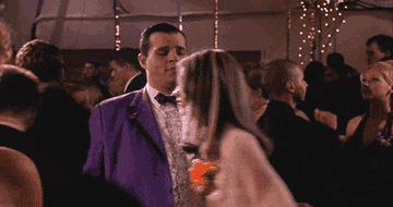 A man in a purple tuxedo, played by the character Mr. Collins from "Mean Girls," is awkwardly dancing at a party full of people