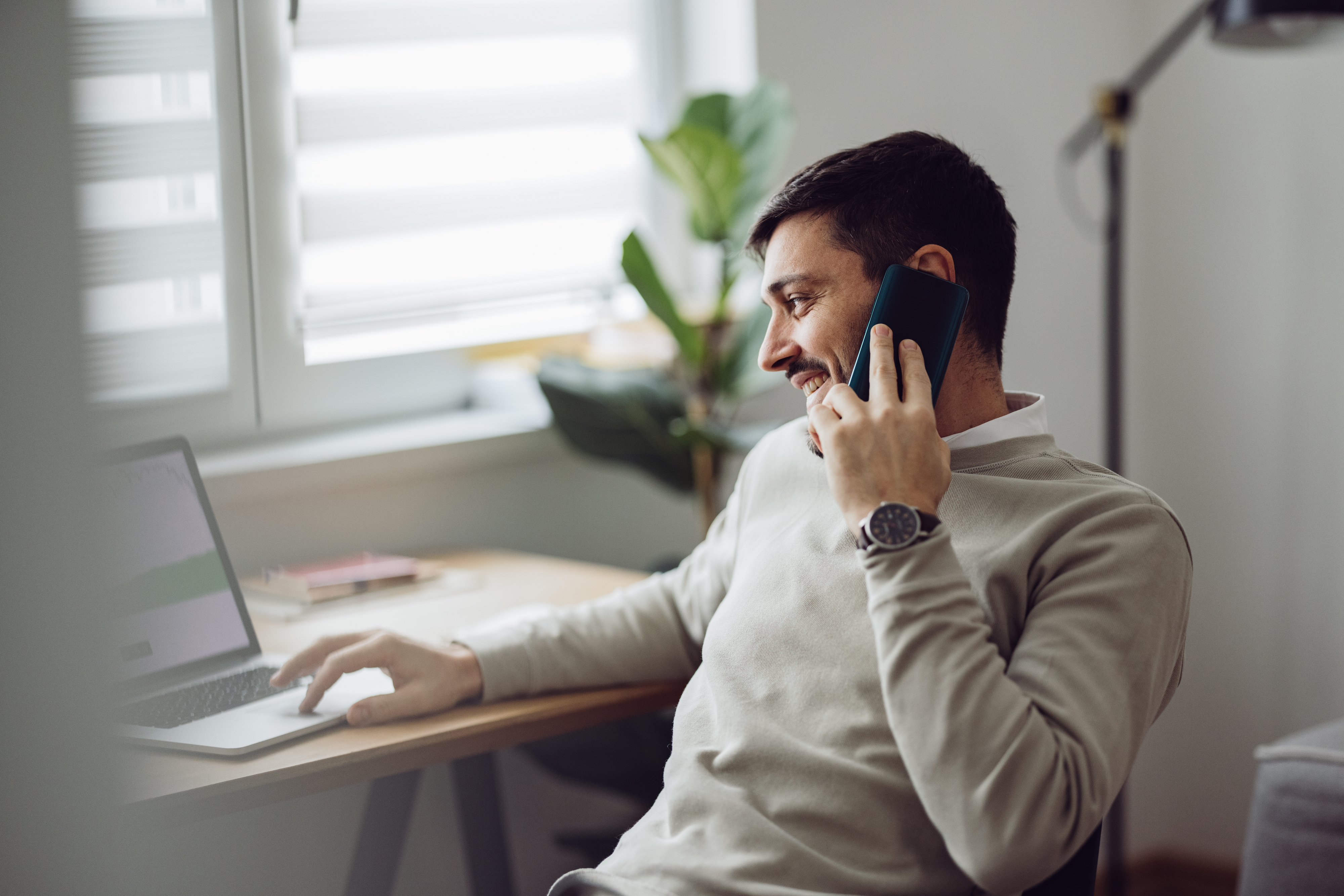 A man is sitting at a desk, smiling while talking on the phone and using a laptop, creating a relaxed and engaging ambiance