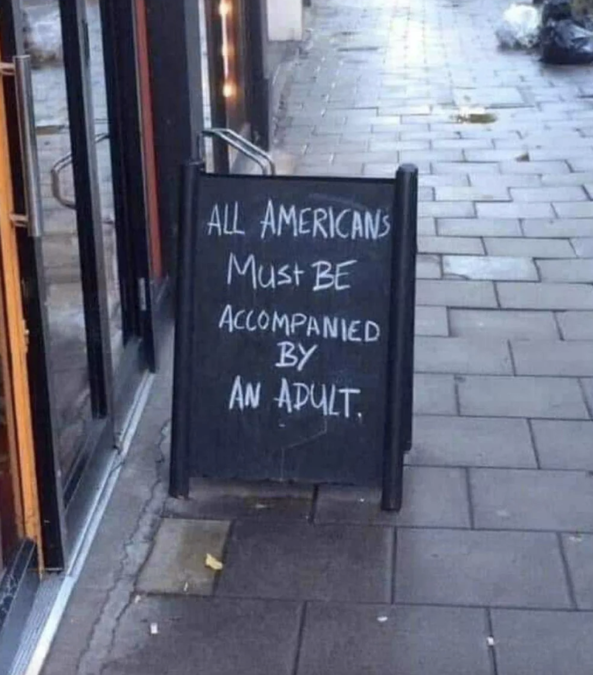 A sidewalk sign outside a building reads: "ALL AMERICANS MUST BE ACCOMPANIED BY AN ADULT."