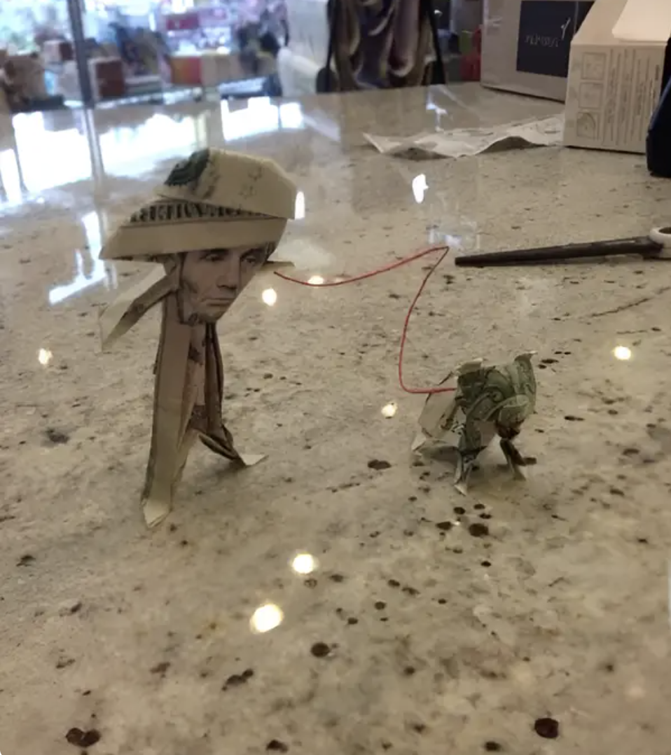 Origami figure wearing a cowboy hat leading an origami frog on a red thread. Both figures are made from folded U.S. dollar bills