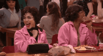 Vanessa Hudgens and Keke Palmer laugh and chat while wearing pink jackets at a table in a lively scene from "Grease: Live."
