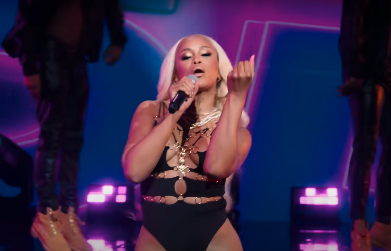 Eve performs on stage, wearing a black, intricately designed bodysuit with gold accessories. She holds a microphone and appears to be mid-song