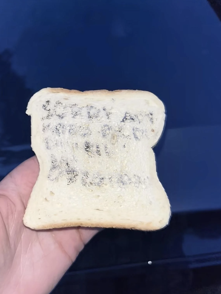 A hand holding a slice of bread with text written on it in what appears to be ink, though the text is difficult to read clearly