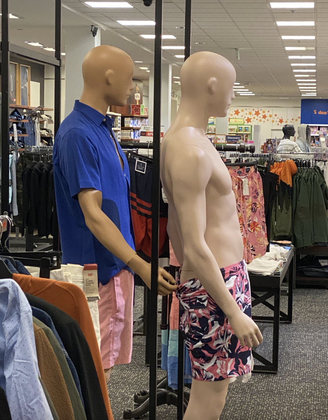 Store display with two mannequins: one wears a short-sleeve shirt and shorts, the other wears only floral-patterned shorts, in a clothing department