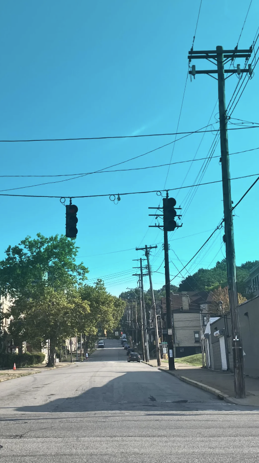 Traffic lights hang over an empty, steep residential street lined with power lines, trees, and houses