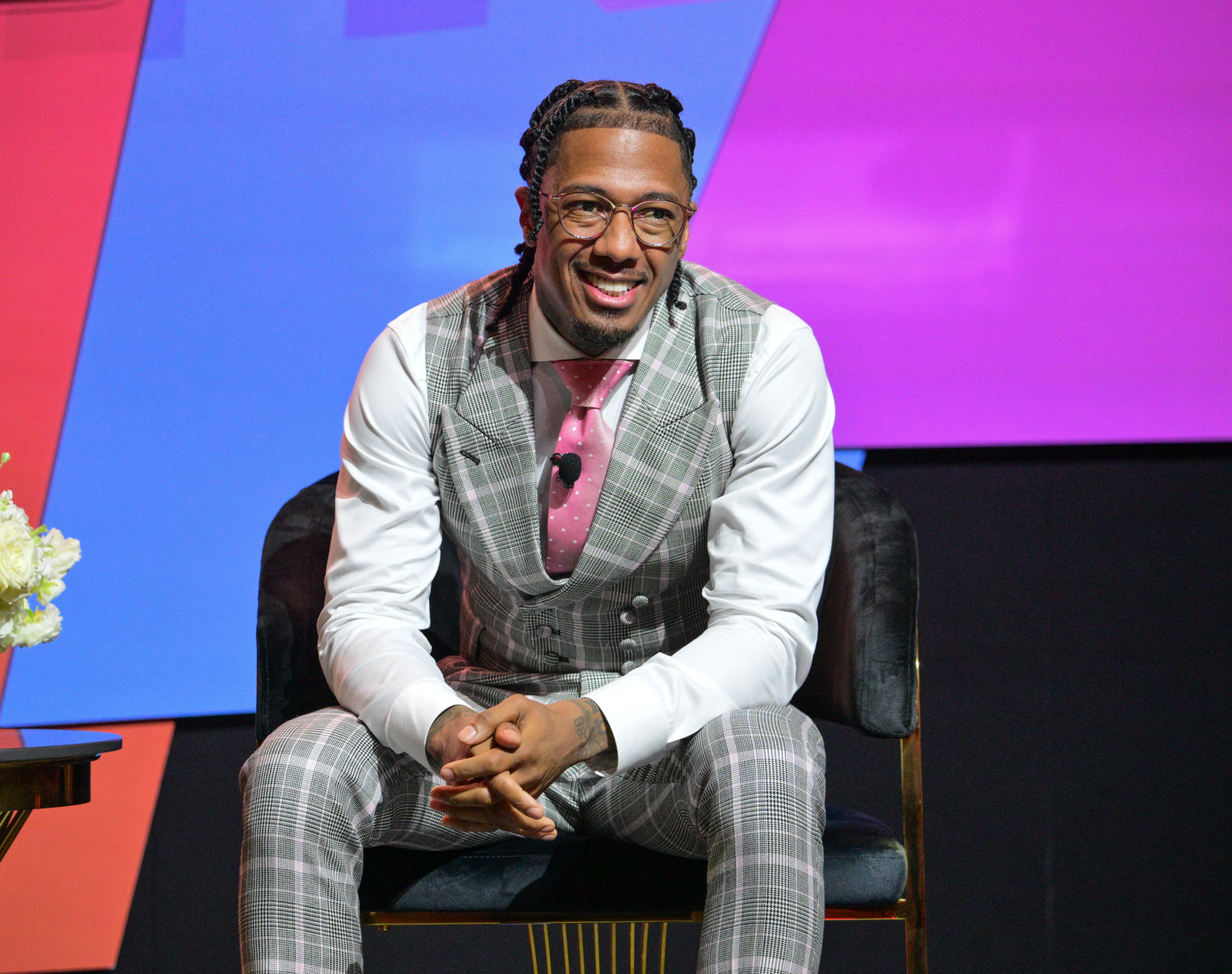 Nick Cannon sits and smiles, wearing a plaid suit with a pink tie and an untucked white shirt