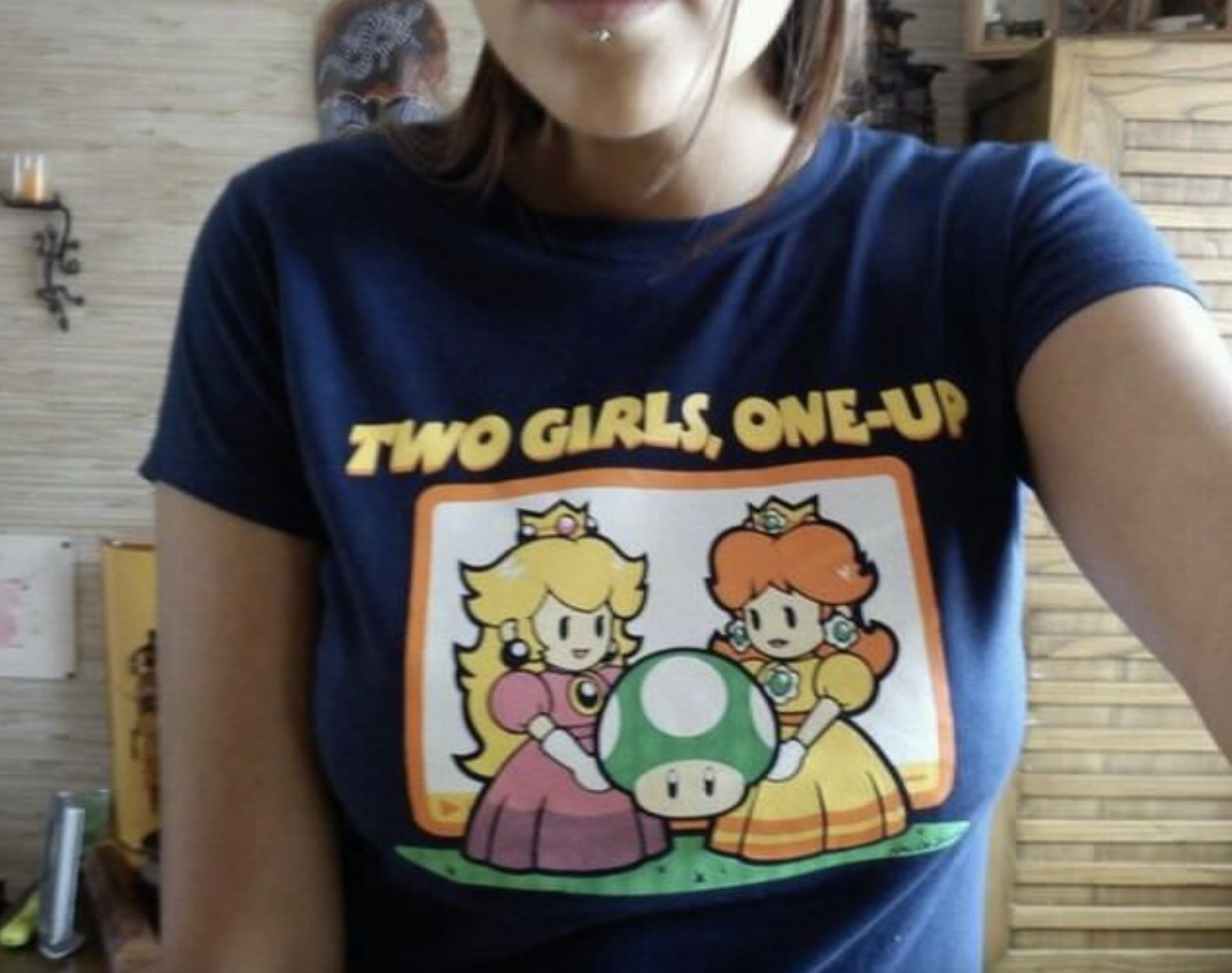 A person wearing a shirt depicting Princess Peach and Princess Daisy from Mario holding a 1-Up mushroom with the text "Two Girls, One-Up."