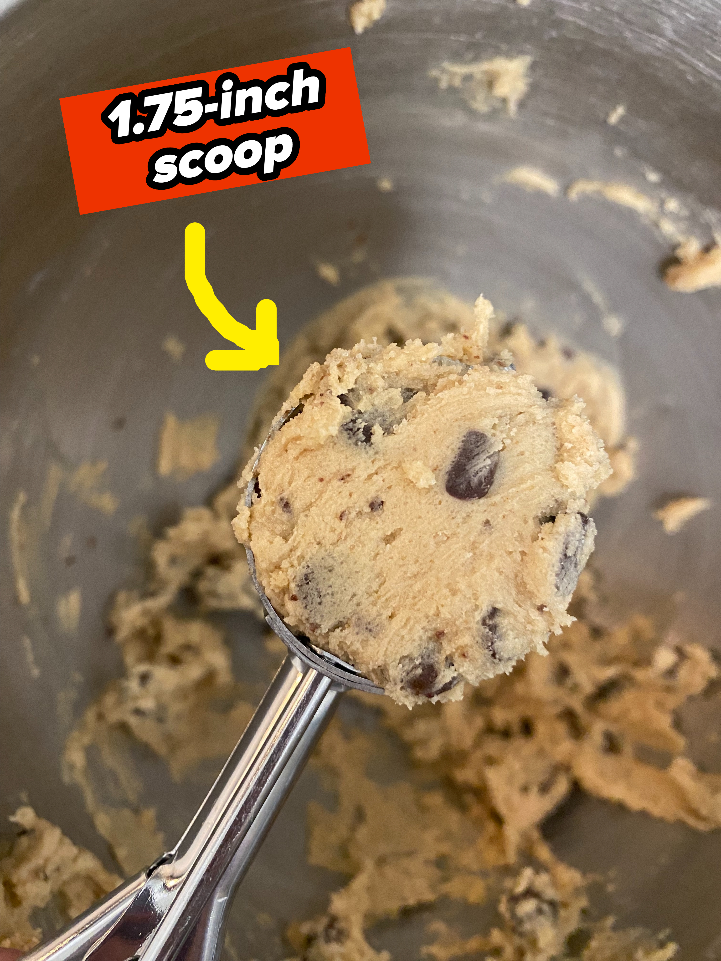 Close-up of a metal cookie scoop filled with chocolate chip cookie dough in a mixing bowl