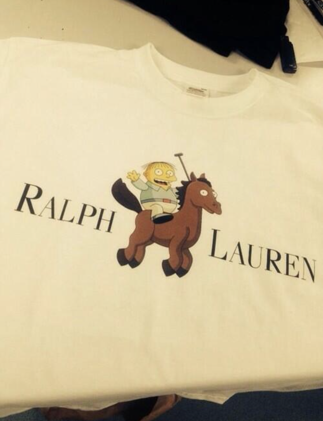 White t-shirt featuring a cartoon character, Ralph Wiggum from The Simpsons, riding a horse with the words "Ralph Lauren" above and below the character