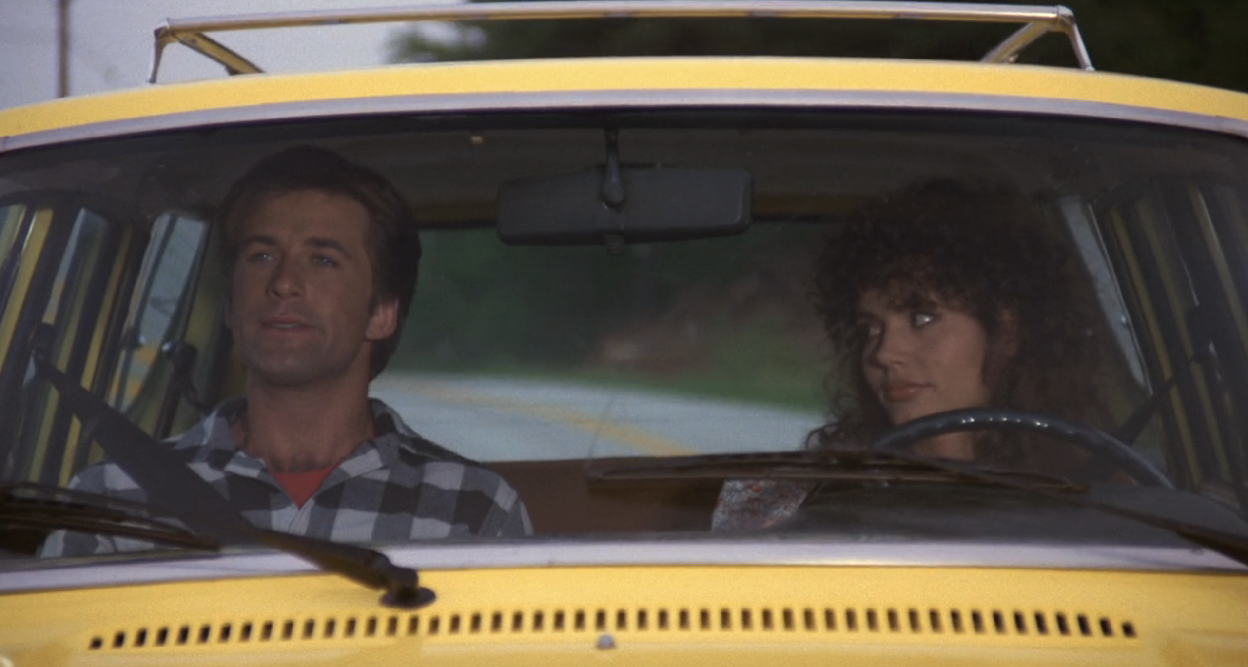 Alec Baldwin and Geena Davis sit in the front seats of a yellow car, the man driving and the woman looking at him. The man wears a checkered shirt. Their expressions seem calm