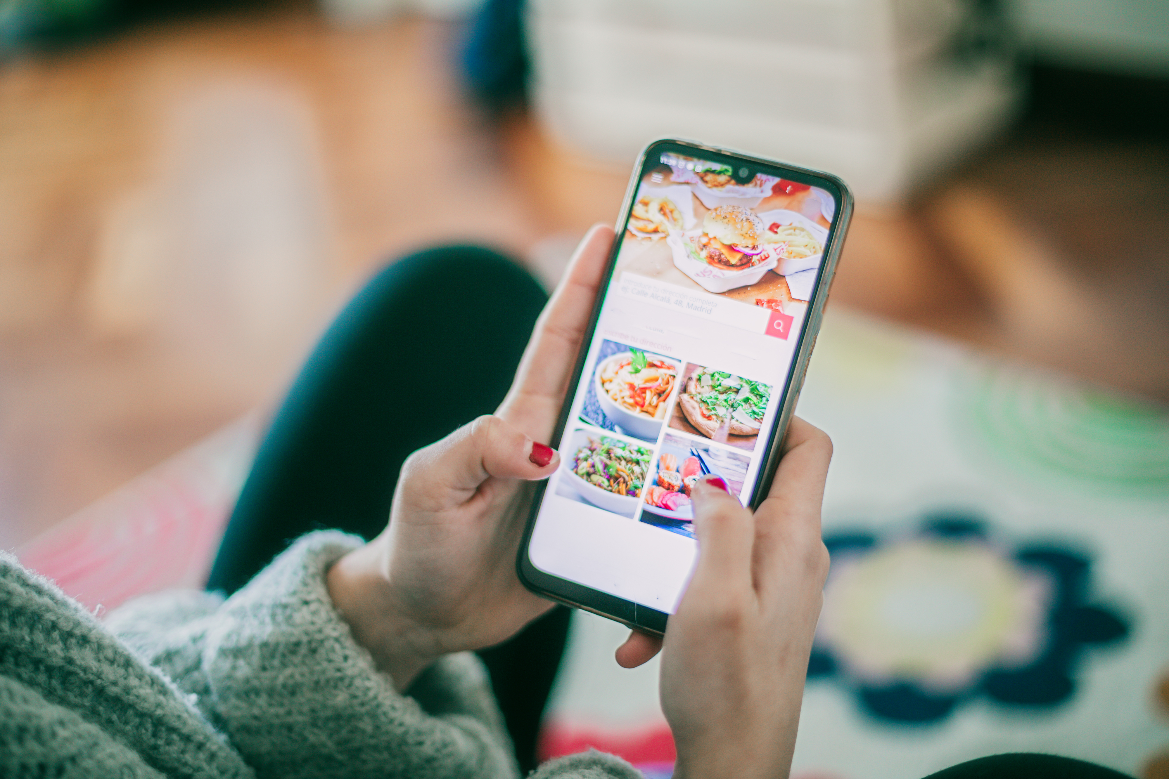 Person using a smartphone to browse a food recipe app with various dish photos. Their hand has red nail polish and they're wearing a cozy sweater