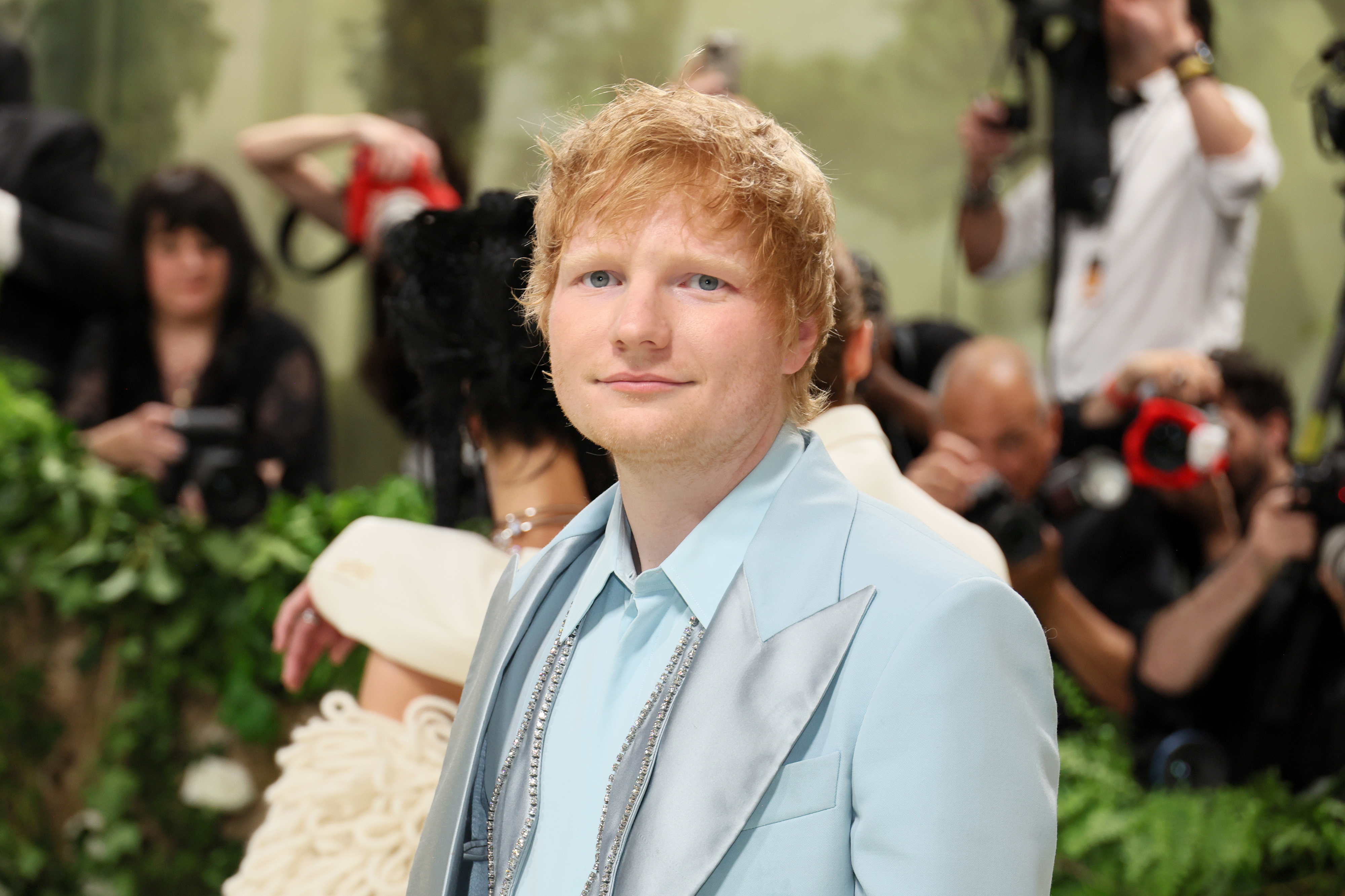Ed Sheeran in a light suit attending a formal event with photographers in the background