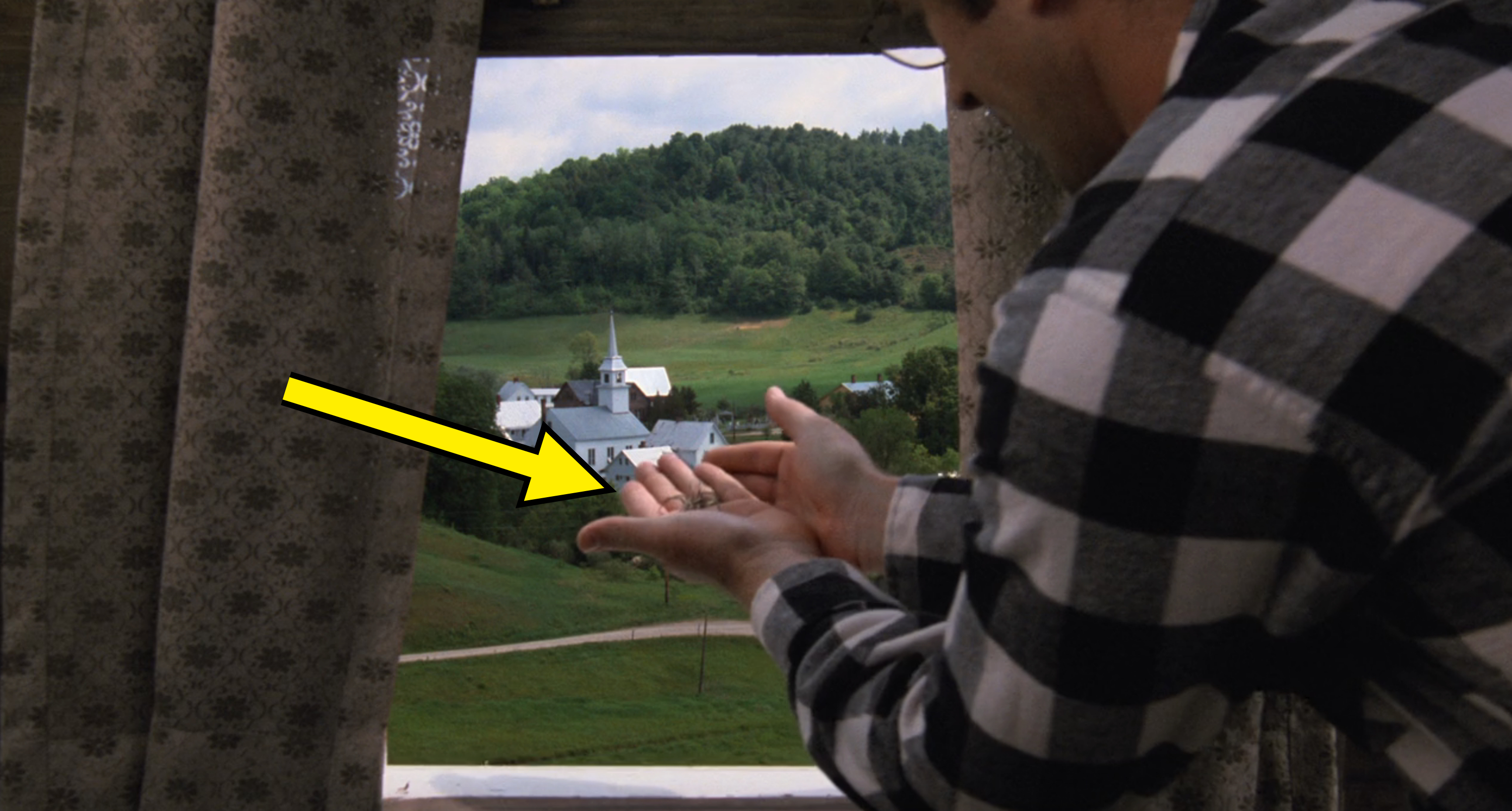 Alec Baldwin in a plaid shirt cups their hands together while looking out a window at a rural landscape with a church and trees in the distance