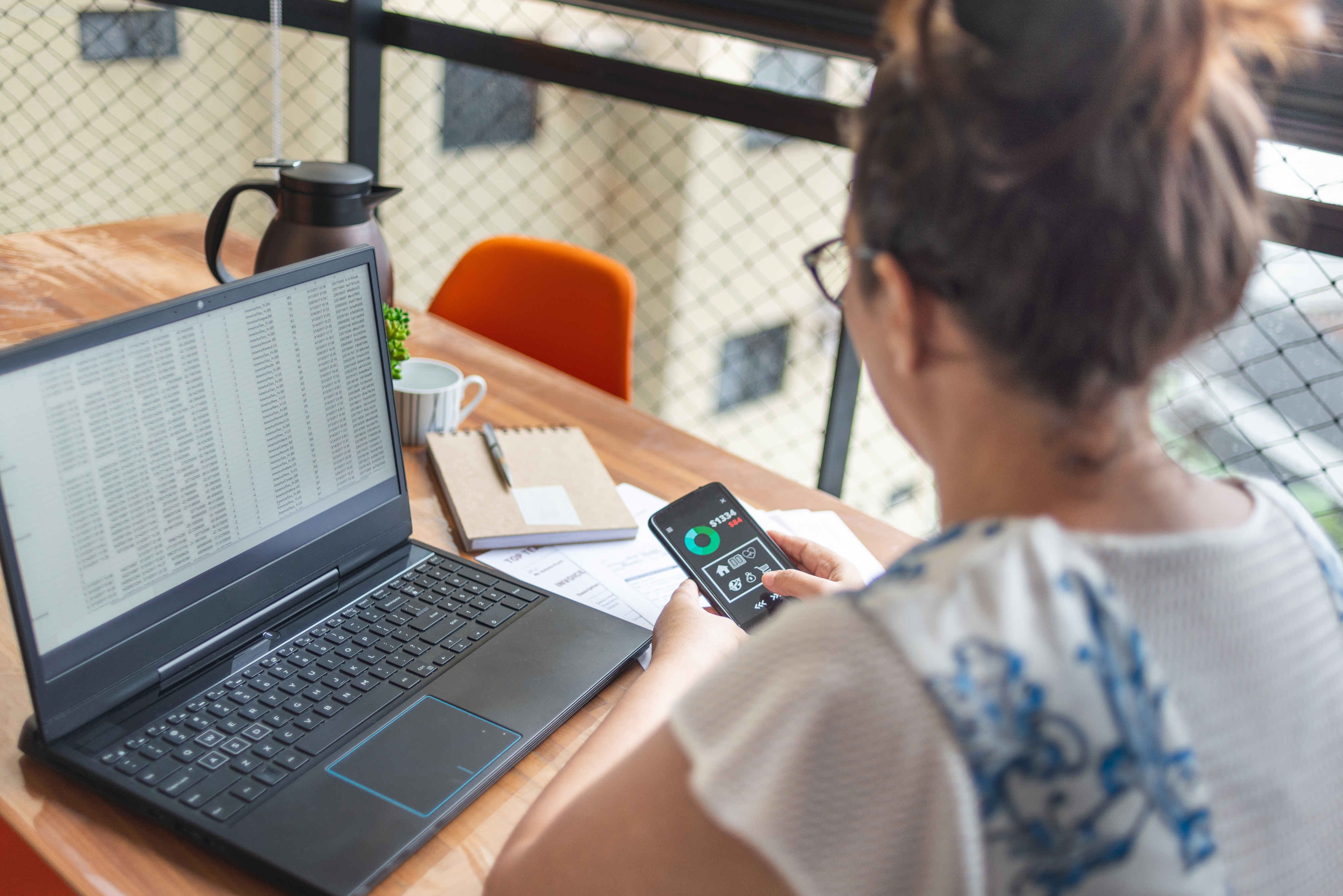 A person with glasses uses a smartphone while sitting at a desk with an open laptop, notepad, coffee mug, and a plant in view