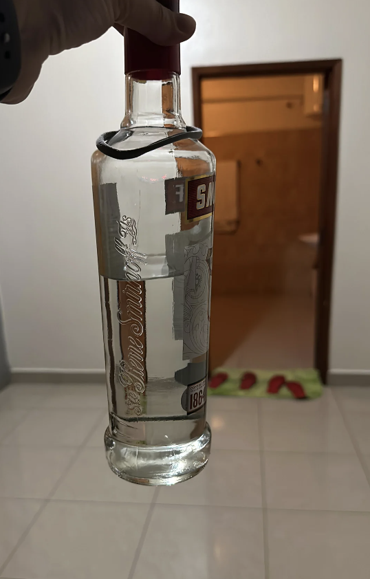 A hand is holding a bottle of Smirnoff vodka in a dimly lit room with a bathroom in the background